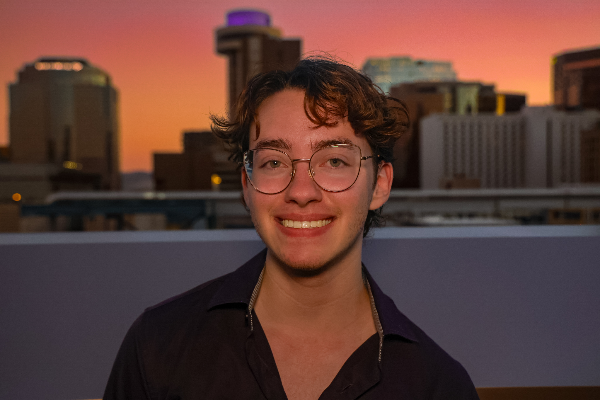 A young man with short brown curly hair and green eyes smiling in front of the downtown Phoenix sunset